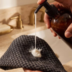 Hand holding a brown bottle with a pump dispensing soap onto a black and white checkered towel over a sink.
